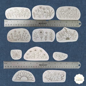K&ouml;nnte beinhalten: Eine Sammlung von Stoffflicken mit floralen Motiven in verschiedenen Formen. Jeder Flicken zeigt ein anderes botanisches Design, darunter Blumen, Bl&auml;tter und ein Schmetterling, alles in einer neutralen Farbe. Ein Lineal liegt zur Gr&ouml;&szlig;enbestimmung &uuml;ber den Flicken.