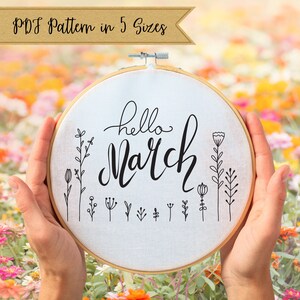 May include: A white embroidery hoop with the text "hello March" in black cursive lettering. The hoop is surrounded by a floral design with small black flowers and stems.