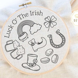 May include: A black and white embroidery hoop with the text "Luck O' The Irish" and various Irish symbols including a pot of gold, a rainbow, a four-leaf clover, a horseshoe, a beer mug, a leprechaun hat, and a shoe.