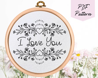 Puede incluir: Un patrón de bordado en blanco y negro de una corona floral con el texto "I Love You" en el centro. El patrón se muestra en un aro de bordado redondo.