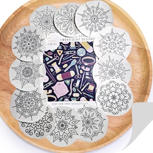 Könnte beinhalten: Ein Set mit 10 runden, silberfarbenen Stick- und Stickdesigns. Die Designs sind filigran und detailliert und zeigen Mandalas und florale Muster. Die Designs sind auf einem Holzteller angeordnet.