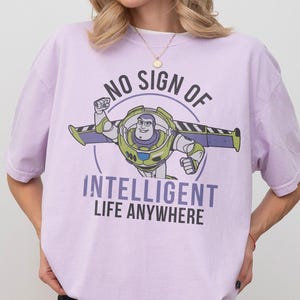 Pu&ograve; includere: T-shirt lilla chiaro con grafica di Buzz Lightyear e la scritta "NO SIGN OF INTELLIGENT LIFE ANYWHERE". La maglietta &egrave; un Comfort Colors Orchid. Il design presenta Buzz Lightyear in posa di volo.