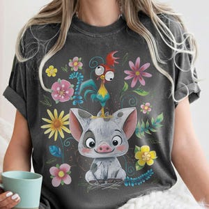 Puede incluir: Camiseta gris oscuro con un cerdo de dibujos animados y un gallo, rodeados de flores coloridas. El dise&ntilde;o incluye elementos florales amarillos, rosas y azules. La camiseta tiene mangas cortas y un corte holgado.