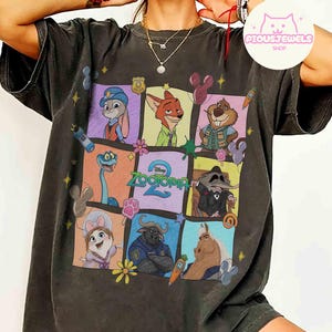 May include: Dark gray t-shirt featuring a colorful graphic of characters from the movie Zootopia. The design includes Judy Hopps, Nick Wilde, and other characters in a grid pattern. The text "Zootopia 2" is also visible.