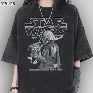 Op de afbeelding: Graphietkleurig Star Wars T-shirt met een afbeelding van The Mandalorian met Grogu. Het Star Wars-logo staat boven de afbeelding. De tekst "I'M A MANDALORIAN. WEAPONS ARE PART OF MY RELIGION." staat eronder.