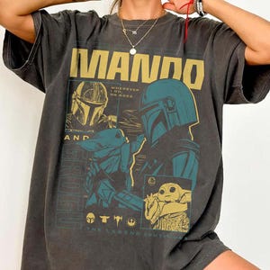May include: Dark gray t-shirt featuring a graphic design with the text "MANDO" in gold letters. The design includes images of a Mandalorian helmet and Grogu, with the text "WHEREVER I GO HE GOES" and "THE LEGEND CONTINUES".