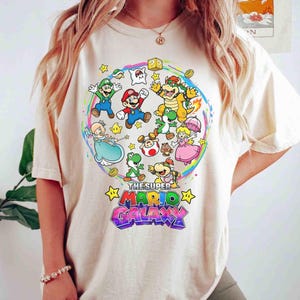 K&ouml;nnte beinhalten: Cremefarbenes T-Shirt mit einem farbenfrohen Grafikdesign von Charakteren aus dem Videospiel Super Mario Galaxy. Das Design zeigt Mario, Luigi, Prinzessin Peach und Bowser, mit dem Text "THE SUPER MARIO GALAXY".