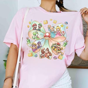Op de afbeelding: Lichtroze t-shirt met een kleurrijke afbeelding van verschillende personages uit de Super Mario-videogameserie. Het ontwerp bevat Mario, Luigi, Princess Peach, Yoshi en Bowser, gerangschikt in een boogvormig frame.