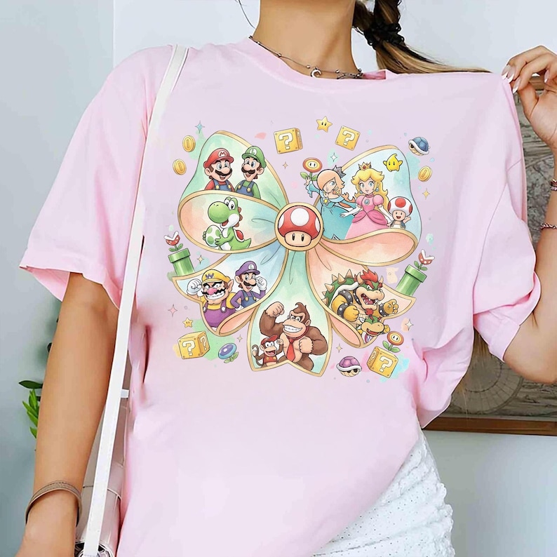 Op de afbeelding: Lichtroze t-shirt met een kleurrijke afbeelding van verschillende personages uit de Super Mario-videogameserie. Het ontwerp bevat Mario, Luigi, Princess Peach, Yoshi en Bowser, gerangschikt in een boogvormig frame.