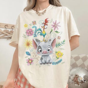 Puede incluir: Camiseta color crema con un gr&aacute;fico colorido de un cerdo, un gallo y flores. El cerdo es gris y blanco con una cara sonriente. El gallo es azul, amarillo y rojo. Las flores son rosas, amarillas y moradas.
