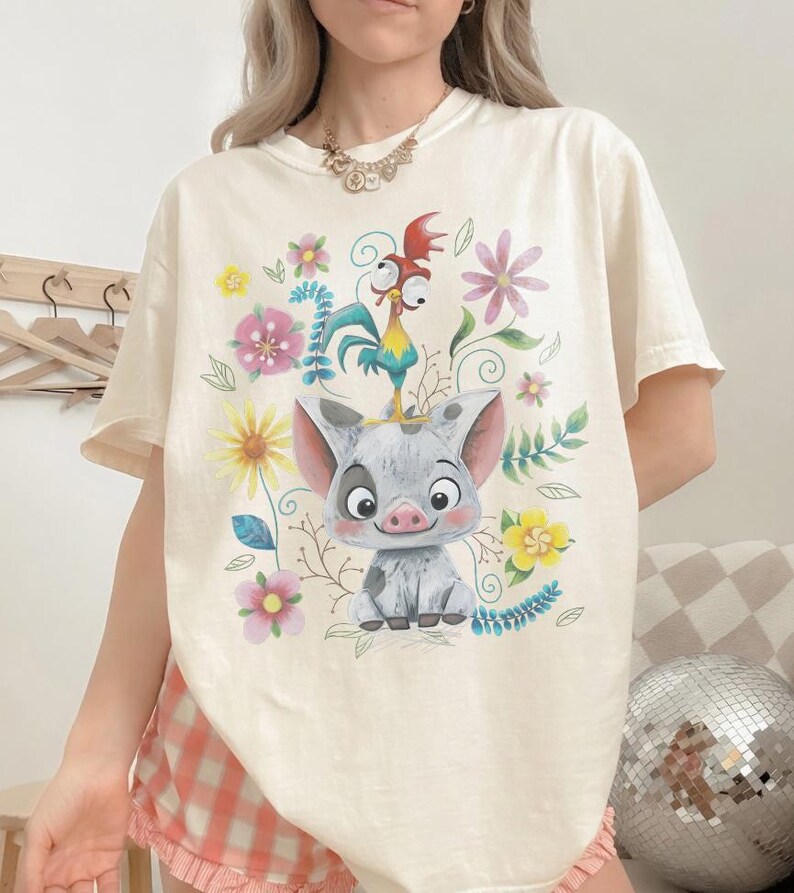 Puede incluir: Camiseta color crema con un gr&aacute;fico colorido de un cerdo, un gallo y flores. El cerdo es gris y blanco con una cara sonriente. El gallo es azul, amarillo y rojo. Las flores son rosas, amarillas y moradas.