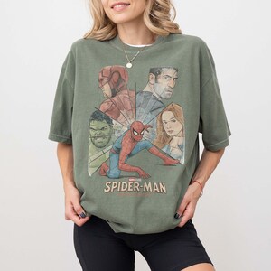 May include: An olive green t-shirt with a graphic featuring Spider-Man and other Marvel characters. The design includes the Hulk, Daredevil, and Captain Marvel. The words "SPIDER-MAN" are printed in white at the bottom of the graphic.