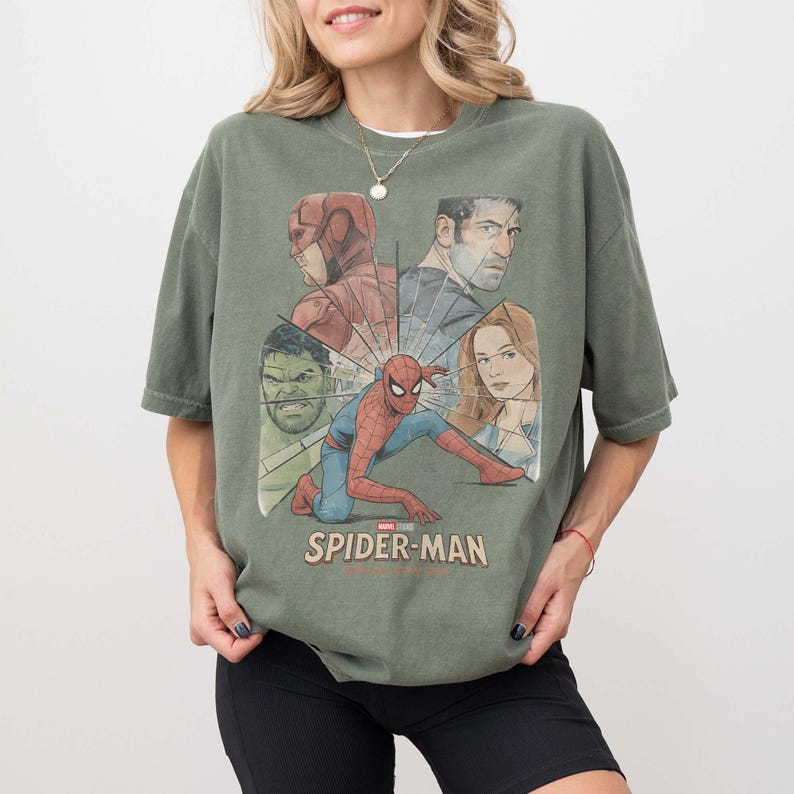 May include: An olive green t-shirt with a graphic featuring Spider-Man and other Marvel characters. The design includes the Hulk, Daredevil, and Captain Marvel. The words "SPIDER-MAN" are printed in white at the bottom of the graphic.