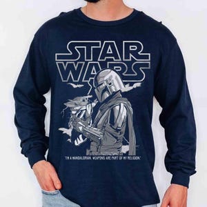 Op de afbeelding: Marineblauwe longsleeve met een Star Wars-ontwerp. Het toont The Mandalorian met Grogu, met de tekst "I'M A MANDALORIAN, WEAPONS ARE PART OF MY RELIGION."
