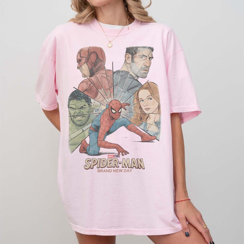 May include: A pale pink t-shirt with a Spider-Man graphic and other Marvel characters. The graphic includes the text "SPIDER-MAN BRAND NEW DAY". The shirt has short sleeves and a crew neck.