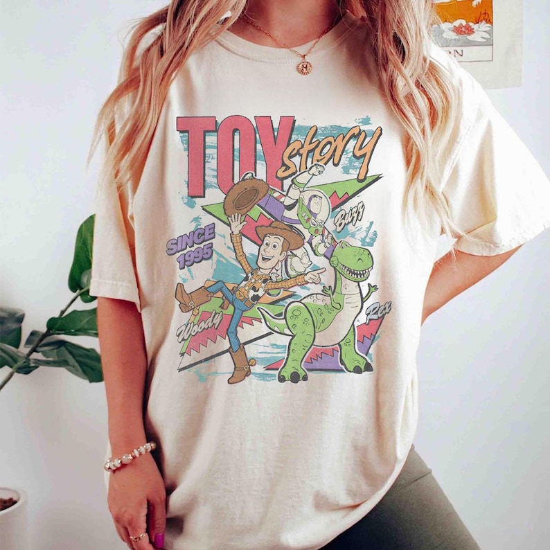 May include: Cream-colored t-shirt featuring a graphic of Toy Story characters Woody, Buzz Lightyear, and Rex, with the text "TOY STORY" and "SINCE 1995". The design includes a retro comic book style with vibrant colors.