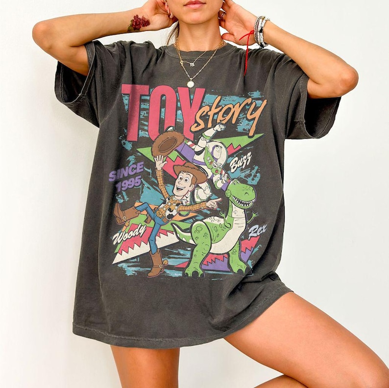 May include: Dark grey t-shirt featuring a colorful graphic of Toy Story characters, including Woody, Buzz Lightyear, and Rex. The text "Toy Story" is displayed above the characters, with "Since 1995" below. The shirt has a vintage, distressed look.