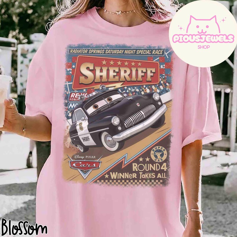 May include: Pink t-shirt featuring a graphic of Sheriff from Disney Pixar's Cars. The design includes the text "RADIATOR SPRINGS SATURDAY NIGHT SPECIAL RACE" and "ROUND 4 WINNER TAKES ALL". The shirt has a crew neck and short sleeves.