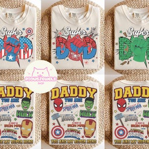 May include: A collection of six cream-colored t-shirts featuring graphic prints of superhero-themed designs. The top row displays the word Super with a fist replacing the letter O, while the bottom row features the text Daddy You Are My Super Hero.