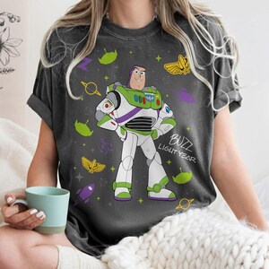 May include: Dark gray t-shirt featuring a cartoon illustration of Buzz Lightyear from Toy Story. The design includes Buzz in his spacesuit, surrounded by space-themed graphics and the text "Buzz Lightyear".