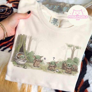 May include: Cream-colored sweatshirt featuring a cartoon illustration of a green alien character in a forest setting. The design includes the character in a pod, playing with a ball, and near a log. The word "COO!" is also visible.