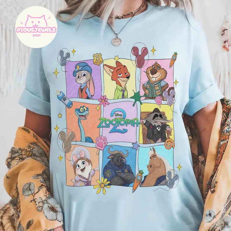 May include: Light blue t-shirt featuring a colorful graphic of characters from the animated movie Zootopia. The design includes Judy Hopps, Nick Wilde, and other characters in a grid pattern. The word "Zootopia" is also visible.