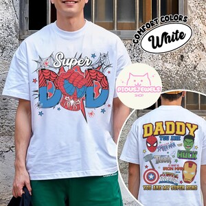 May include: A white t-shirt featuring a front graphic with the text Super DAD and a spider-web motif. The back graphic displays the text DADDY YOU ARE and lists various superhero characters including Spider-Man, Hulk, Thor, and Iron Man.