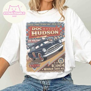 May include: White t-shirt featuring a graphic of Doc Hudson from the Disney Pixar Cars movie. The design includes the text "Radiator Springs Saturday Night Special Race" and "Winner Takes All". The car is dark blue with the number 51.