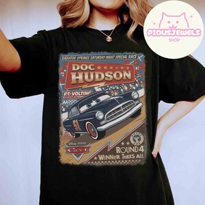 May include: Black t-shirt featuring a vintage-style graphic of Doc Hudson from the Disney Pixar Cars movie. The design includes the text "RADIATOR SPRINGS SATURDAY NIGHT SPECIAL RACE" and "ROUND 4 WINNER TAKES ALL".