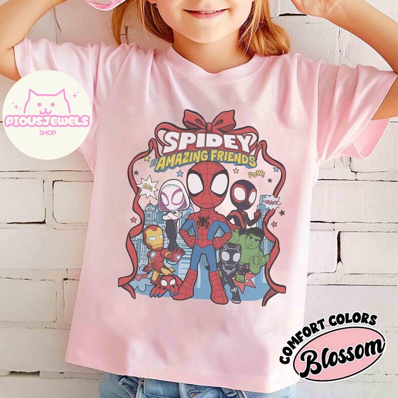 Puede incluir: Una camiseta rosa claro con un estampado de Spidey y sus Incre&iacute;bles Amigos. El dise&ntilde;o incluye varios personajes de dibujos animados, el texto "Spidey Amazing Friends" y la palabra "Pow!". La camiseta tambi&eacute;n tiene el texto "Comfort Colors Blossom".