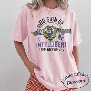 Pu&ograve; includere: T-shirt rosa chiaro con una grafica di Buzz Lightyear e la scritta "NO SIGN OF INTELLIGENT LIFE ANYWHERE". La maglietta ha anche le parole "Comfort Colors Blossom" stampate.