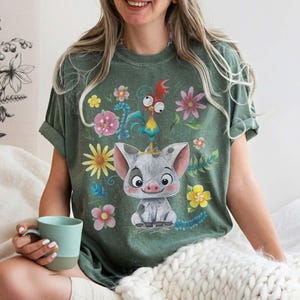 Puede incluir: Camiseta verde con un cerdo de dibujos animados con una corona, rodeado de flores coloridas y un gallo. La camiseta tiene un corte relajado, ideal para el uso diario. El dise&ntilde;o es brillante y atractivo, para los fan&aacute;ticos de los personajes animados.
