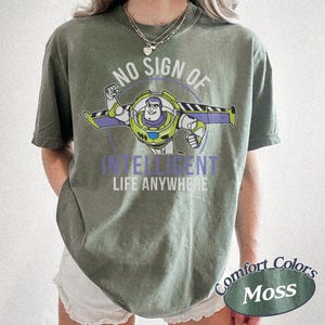 Pu&ograve; includere: T-shirt verde muschio con grafica di Buzz Lightyear e la scritta "NO SIGN OF INTELLIGENT LIFE ANYWHERE". Il design include un bordo circolare e il logo Comfort Colors.