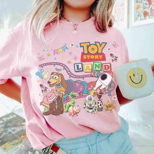 May include: Pale pink t-shirt with a vibrant "Toy Story Land" design, showcasing characters and imagery from the film. The graphic includes a dog, dinosaur, rocket, and the words "Toy Story Land". A white mug with a yellow smiley face is also visible.