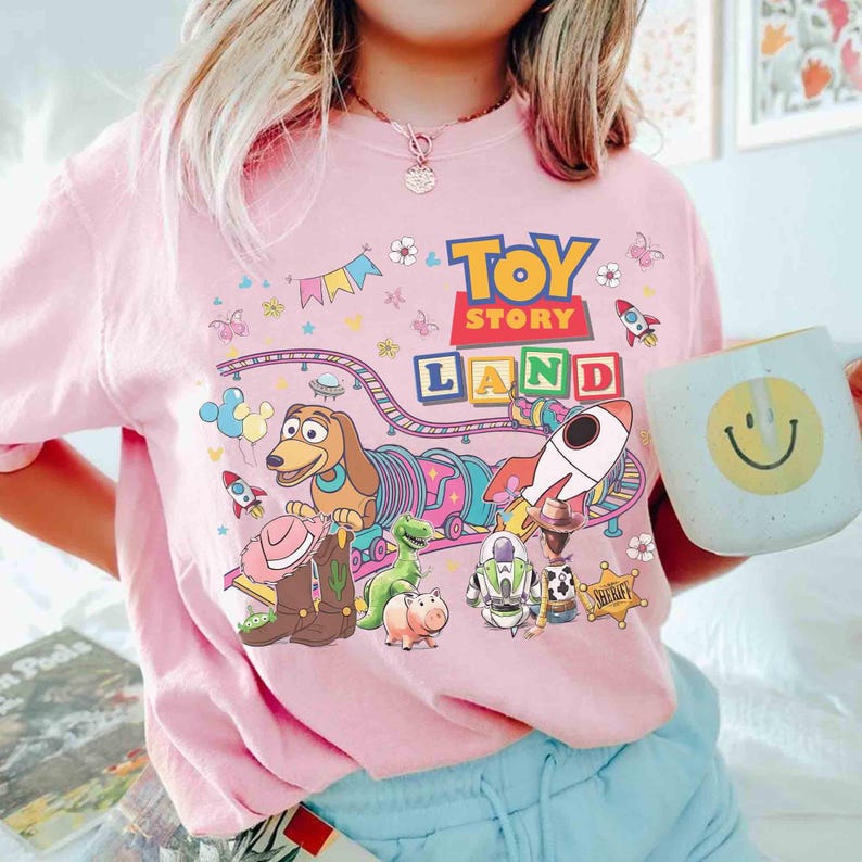 May include: Pale pink t-shirt with a vibrant "Toy Story Land" design, showcasing characters and imagery from the film. The graphic includes a dog, dinosaur, rocket, and the words "Toy Story Land". A white mug with a yellow smiley face is also visible.
