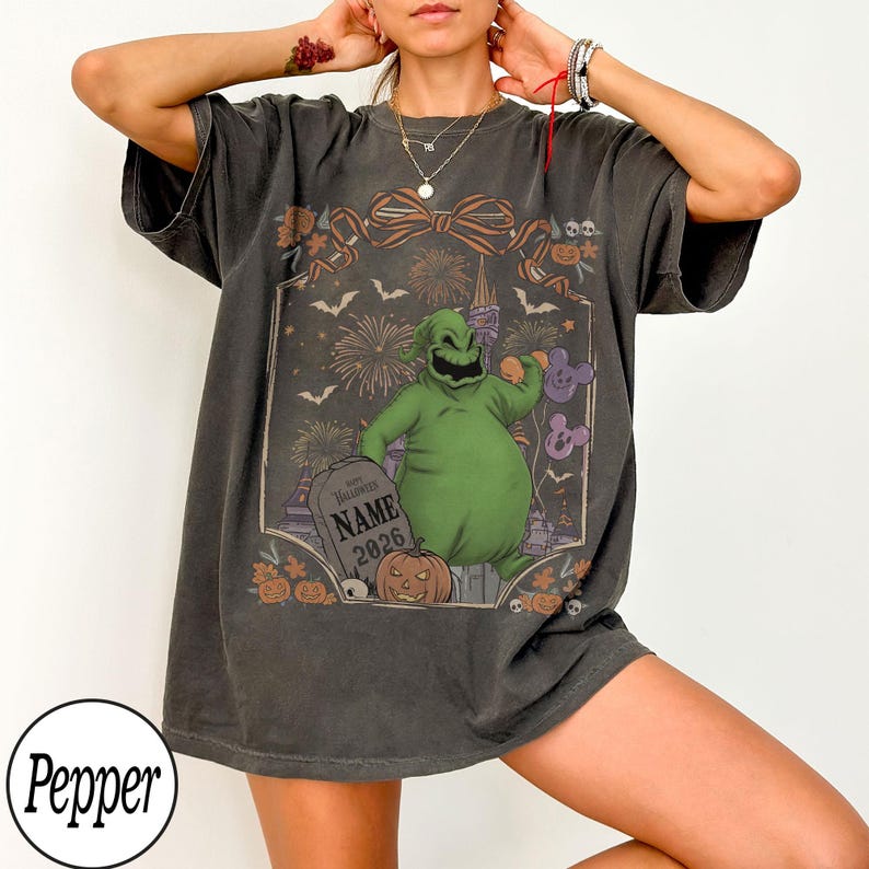 K&ouml;nnte beinhalten: Dunkelgraues T-Shirt mit einem gr&uuml;nen Oogie Boogie-Charakterdesign, einem Grabstein mit der Aufschrift "NAME 2026", Feuerwerk, Flederm&auml;usen und K&uuml;rbissen. Das Design wird von einer Schleife umrahmt. Das T-Shirt hat kurze &Auml;rmel.