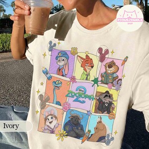 May include: Ivory-colored t-shirt featuring a colorful graphic of characters from the Disney movie "Zootopia." The design includes Judy Hopps, Nick Wilde, and other characters in a grid pattern. The shirt has a crew neck and short sleeves.
