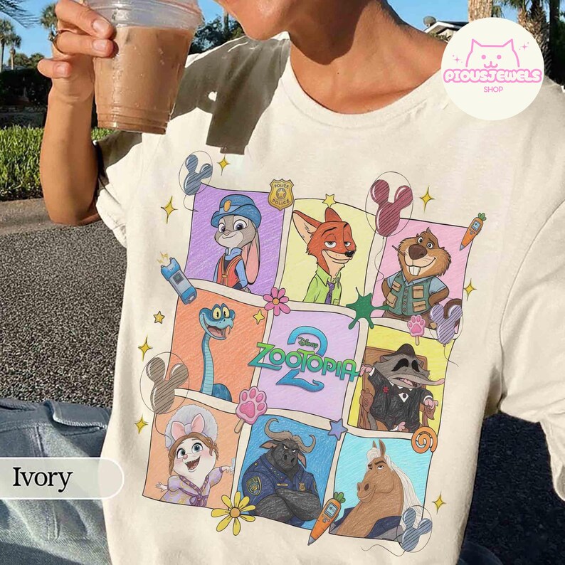 May include: Ivory-colored t-shirt featuring a colorful graphic of characters from the Disney movie "Zootopia." The design includes Judy Hopps, Nick Wilde, and other characters in a grid pattern. The shirt has a crew neck and short sleeves.