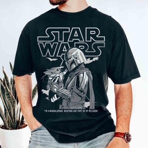 Op de afbeelding: Zwart T-shirt met een Star Wars-afbeelding. Het ontwerp bevat het Star Wars-logo, een illustratie van The Mandalorian met Grogu en de quote: "IK BEN EEN MANDALORIAN. WAPENS ZIJN ONDERDEEL VAN MIJN RELIGIE."