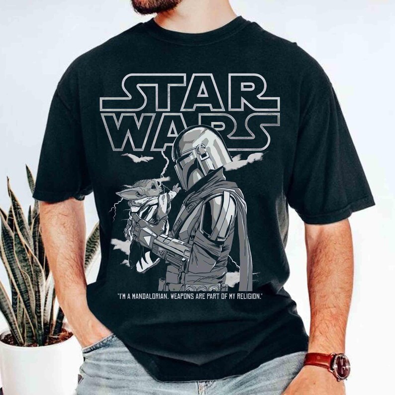 Op de afbeelding: Zwart T-shirt met een Star Wars-afbeelding. Het ontwerp bevat het Star Wars-logo, een illustratie van The Mandalorian met Grogu en de quote: "IK BEN EEN MANDALORIAN. WAPENS ZIJN ONDERDEEL VAN MIJN RELIGIE."