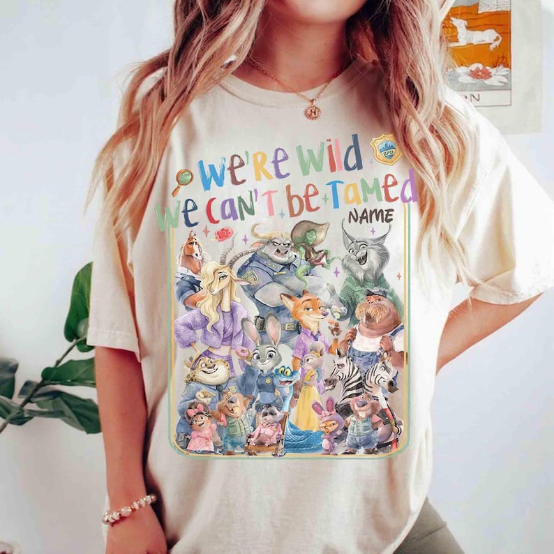Puede incluir: Camiseta color crema con un gr&aacute;fico colorido de varios animales de dibujos animados. El texto "WE'RE WILD WE CAN'T BE TAMED" se muestra encima de los personajes. Se incluye un espacio para un nombre. El dise&ntilde;o est&aacute; delineado en verde.
