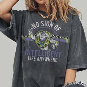 Pu&ograve; includere: T-shirt grigio scuro con una grafica di Buzz Lightyear e la scritta "NO SIGN OF INTELLIGENT LIFE ANYWHERE". La maglietta &egrave; una Comfort Colors Pepper.