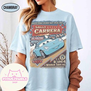May include: Light blue t-shirt featuring a graphic of Sally Carrera from the Disney Pixar Cars movie. The design includes text that reads "Radiator Springs Saturday Night Special Race" and "Winner Takes All."