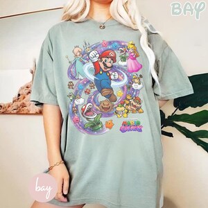 K&ouml;nnte beinhalten: Salbeigr&uuml;nes T-Shirt mit einer farbenfrohen Grafik von Mario aus dem Videospiel Super Mario Galaxy. Das Design zeigt Mario, Prinzessin Peach und andere Charaktere, umgeben von einem wirbelnden Galaxiemuster. Der Text "Mario Galaxy" ist ebenfalls sichtbar.