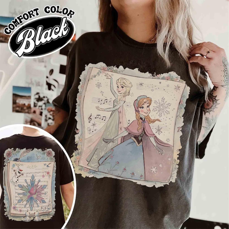 Pode incluir: Camiseta preta Comfort Color com um gr&aacute;fico de duas princesas animadas, notas musicais e detalhes florais. A camisa tamb&eacute;m tem uma imagem menor na parte inferior esquerda das costas com um floco de neve e o texto "Let it go".