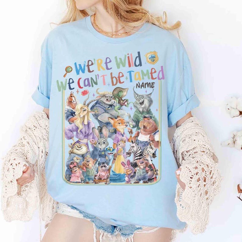 Puede incluir: Camiseta azul claro con un gr&aacute;fico colorido de varios animales de dibujos animados. La camiseta tiene el texto "we're wild we can't be tamed" con la palabra "NAME" debajo. El dise&ntilde;o incluye una insignia y una lupa.