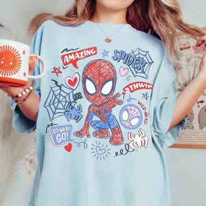 May include: A light blue t-shirt featuring a central graphic of a red and blue cartoon spider character in a crouching pose. The design includes spider webs, hearts, stars, and text that reads Amazing, Spider, Thwip, Web-Head, and Go-Webs-Go.