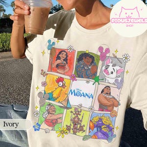 Pu&ograve; includere: T-shirt color avorio con una grafica colorata dei personaggi del film Disney Oceania. Il design include illustrazioni di Oceania, Maui, Pua, Hei Hei e altri, con il logo del film al centro. La maglietta &egrave; decorata con fiori e stelle.
