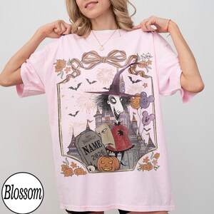 K&ouml;nnte beinhalten: Hellrosa T-Shirt mit einem Halloween-Motiv. Das Design zeigt eine Figur mit Hexenhut, ein Schloss, Flederm&auml;use, K&uuml;rbisse und den Text "Happy Halloween NAME 2026". Das T-Shirt hat eine lockere Passform.