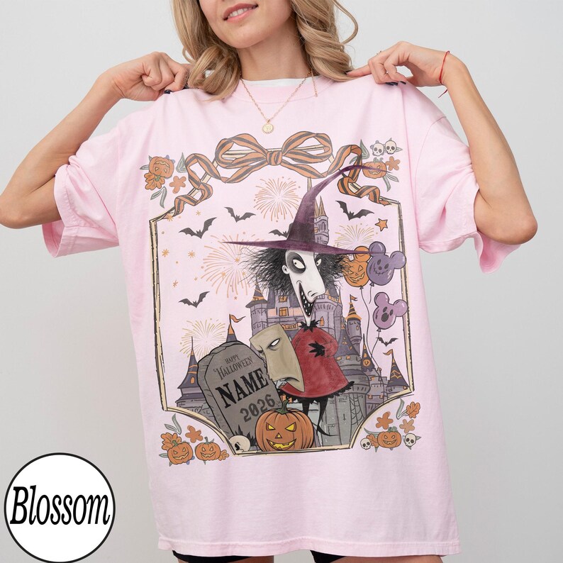 K&ouml;nnte beinhalten: Hellrosa T-Shirt mit einem Halloween-Motiv. Das Design zeigt eine Figur mit Hexenhut, ein Schloss, Flederm&auml;use, K&uuml;rbisse und den Text "Happy Halloween NAME 2026". Das T-Shirt hat eine lockere Passform.